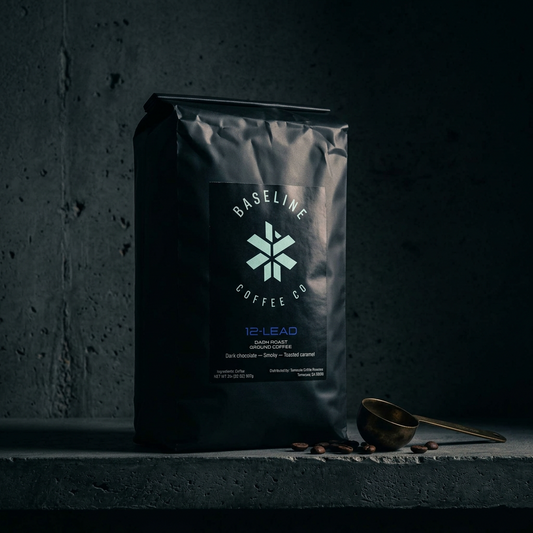 A matte black bag of Baseline Coffee Co. 12-Lead dark chocolate coffee stands on a dark stone surface with scattered coffee beans and a small brass scoop nearby.